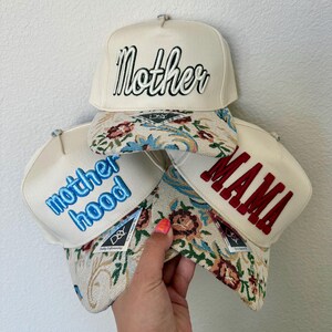 Puede incluir: Cuatro gorras de b&eacute;isbol color crema con texto bordado. Una gorra dice "Mother" en escritura negra, otra "MAMA" en rojo, y una tercera "motherhood" en azul. Las viseras presentan un dise&ntilde;o de tapiz floral.