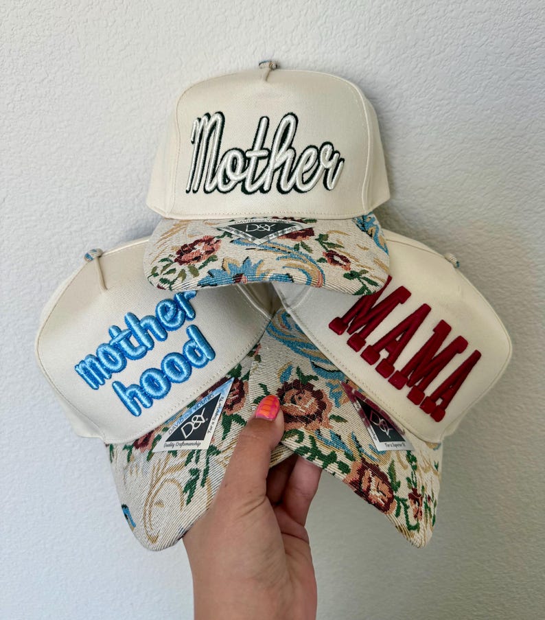 Puede incluir: Cuatro gorras de b&eacute;isbol color crema con texto bordado. Una gorra dice "Mother" en escritura negra, otra "MAMA" en rojo, y una tercera "motherhood" en azul. Las viseras presentan un dise&ntilde;o de tapiz floral.