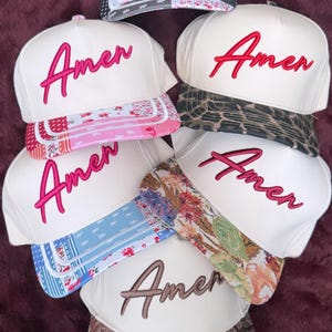 May include: Six baseball caps with white crowns and varied patterned brims. Five caps feature the word Amen embroidered in pink, while the bottom cap features the word Amen in brown. The bottom cap also displays a D&Y brand sticker on the brim.