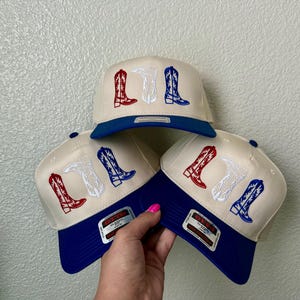 May include: Three baseball caps with a white and blue design. Each cap has an embroidered design of three cowboy boots in red, white, and blue.