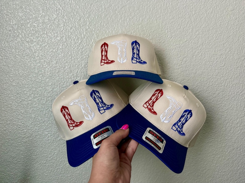 May include: Three baseball caps with a white and blue design. Each cap has an embroidered design of three cowboy boots in red, white, and blue.