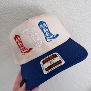 May include: A white and blue baseball cap with a navy blue brim. The front of the cap features an embroidered design of three cowboy boots in red, white, and blue. The cap has a label that reads "OTTO Classic Fit OSFM Mid Profile Maximum Comfort".