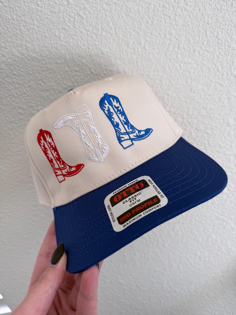 May include: A white and blue baseball cap with a navy blue brim. The front of the cap features an embroidered design of three cowboy boots in red, white, and blue. The cap has a label that reads "OTTO Classic Fit OSFM Mid Profile Maximum Comfort".