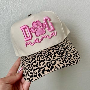 May include: A beige baseball cap with a leopard print brim. The cap features the embroidered text "Dog Mama" in pink, with a paw print design in the center of the text.