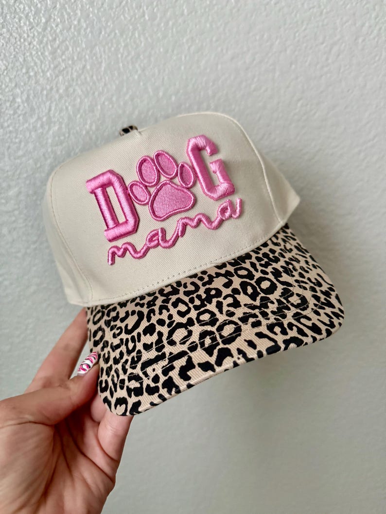 May include: A beige baseball cap with a leopard print brim. The cap features the embroidered text "Dog Mama" in pink, with a paw print design in the center of the text.