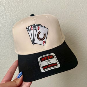 May include: A beige and black baseball cap with an embroidered design featuring playing cards and a horseshoe. The cap has a black brim and the text "OTTO" on a tag.