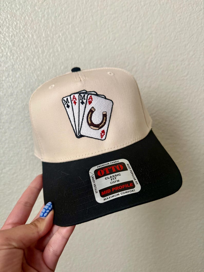 May include: A beige and black baseball cap with an embroidered design featuring playing cards and a horseshoe. The cap has a black brim and the text "OTTO" on a tag.