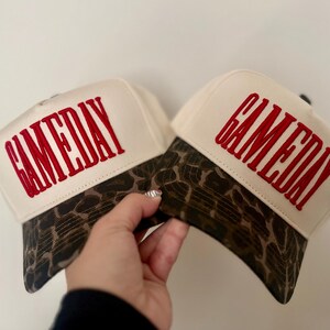 May include: Two cream-colored baseball caps featuring a brown and black leopard print pattern on the brim. Each cap has the word GAMEDAY embroidered in bold red capital letters across the front panel, with a structured crown and curved visor.