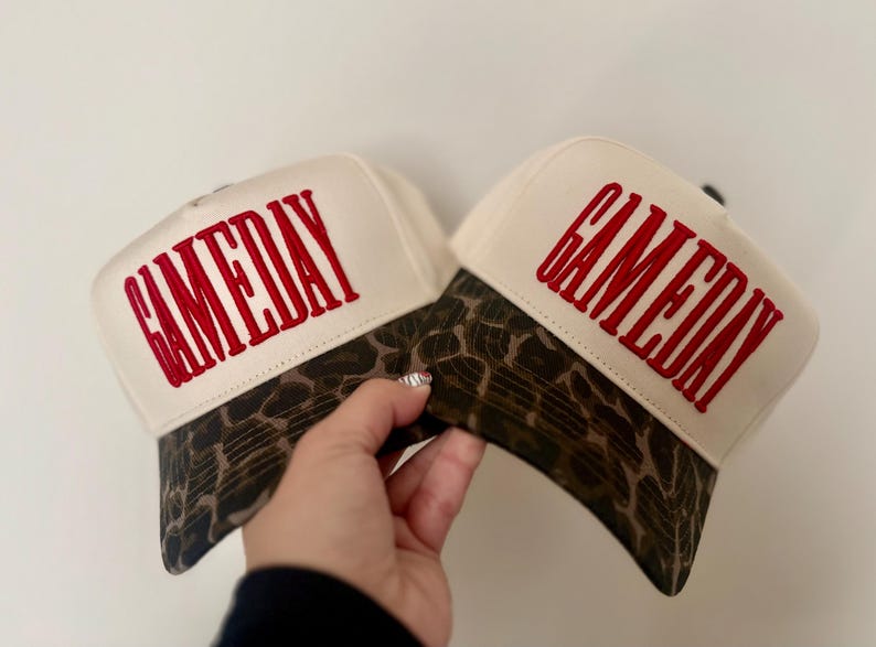 May include: Two cream-colored baseball caps featuring a brown and black leopard print pattern on the brim. Each cap has the word GAMEDAY embroidered in bold red capital letters across the front panel, with a structured crown and curved visor.