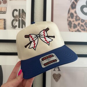 May include: Cream-colored baseball cap with a blue brim. The cap features an embroidered design of a baseball bow with red stitching and brown baseball bats. The cap has a classic fit and mid profile.