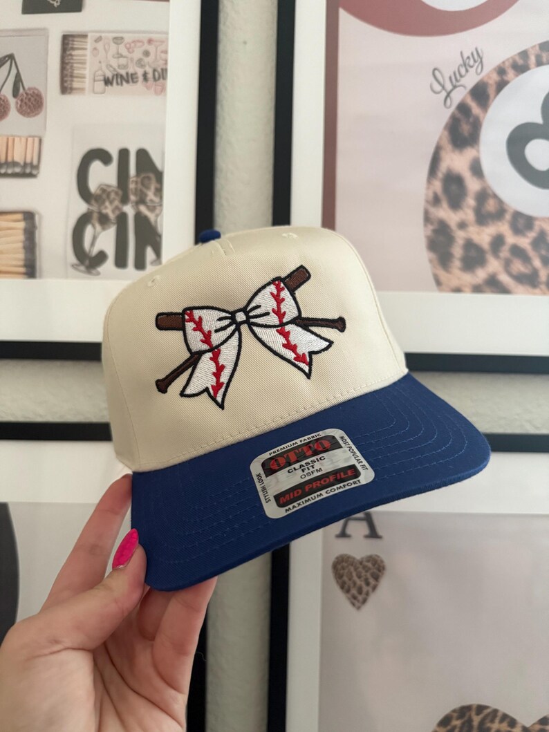 May include: Cream-colored baseball cap with a blue brim. The cap features an embroidered design of a baseball bow with red stitching and brown baseball bats. The cap has a classic fit and mid profile.