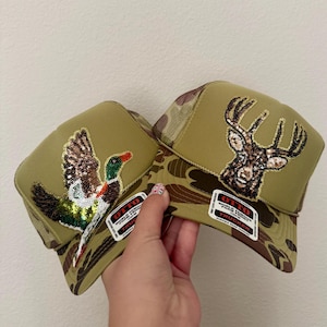 May include: Two olive green trucker hats with camouflage visors. One hat features a sequined duck, the other a sequined deer. Both hats have the text "OTTO WORLD FAMOUS TRUCKER" on a white tag.