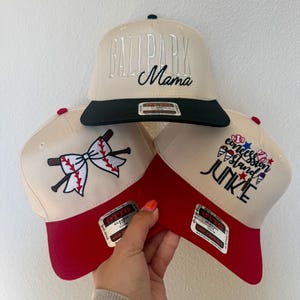May include: Three baseball caps with cream-colored crowns and red brims are displayed. One cap has "BALLPARK Mama" embroidered in silver, another has a baseball and bow design, and the third says "Concession Island Junkie".