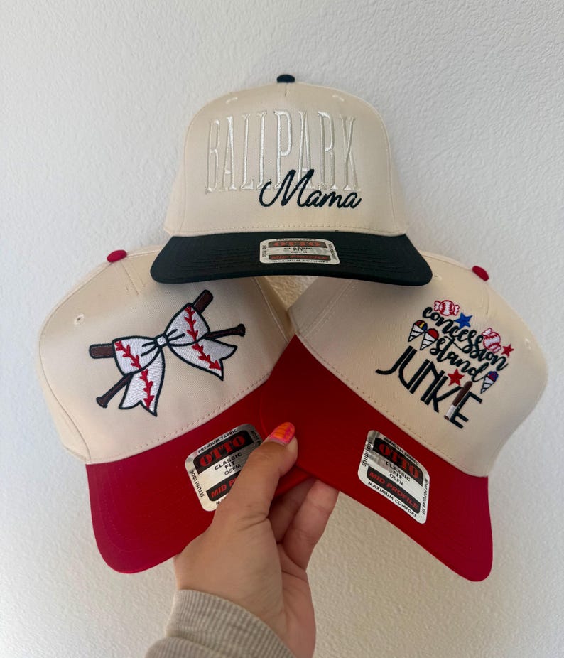 May include: Three baseball caps with cream-colored crowns and red brims are displayed. One cap has "BALLPARK Mama" embroidered in silver, another has a baseball and bow design, and the third says "Concession Island Junkie".