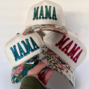 Puede incluir: Tres gorras de camionero blancas con la palabra "MAMA" bordada en verde, turquesa y rojo. Las viseras presentan un dise&ntilde;o de tapiz floral. Las gorras est&aacute;n apiladas, una de ellas sostenida por una mano.