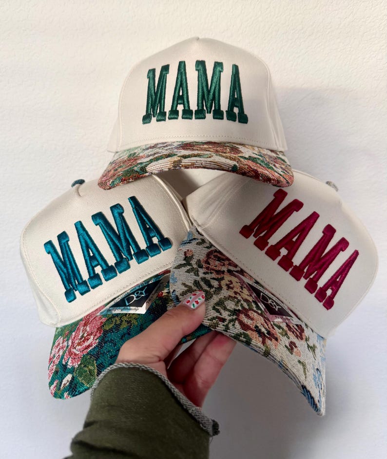 Puede incluir: Tres gorras de camionero blancas con la palabra "MAMA" bordada en verde, turquesa y rojo. Las viseras presentan un dise&ntilde;o de tapiz floral. Las gorras est&aacute;n apiladas, una de ellas sostenida por una mano.