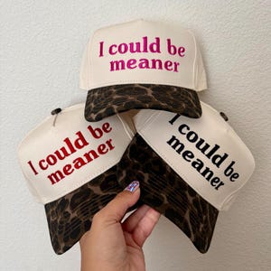 May include: Three trucker hats with cream-colored front panels and brown leopard print brims. Each hat features the embroidered text "I could be meaner" in different colors: one in hot pink, one in red, and one in black, displayed in a stacked arrangement.