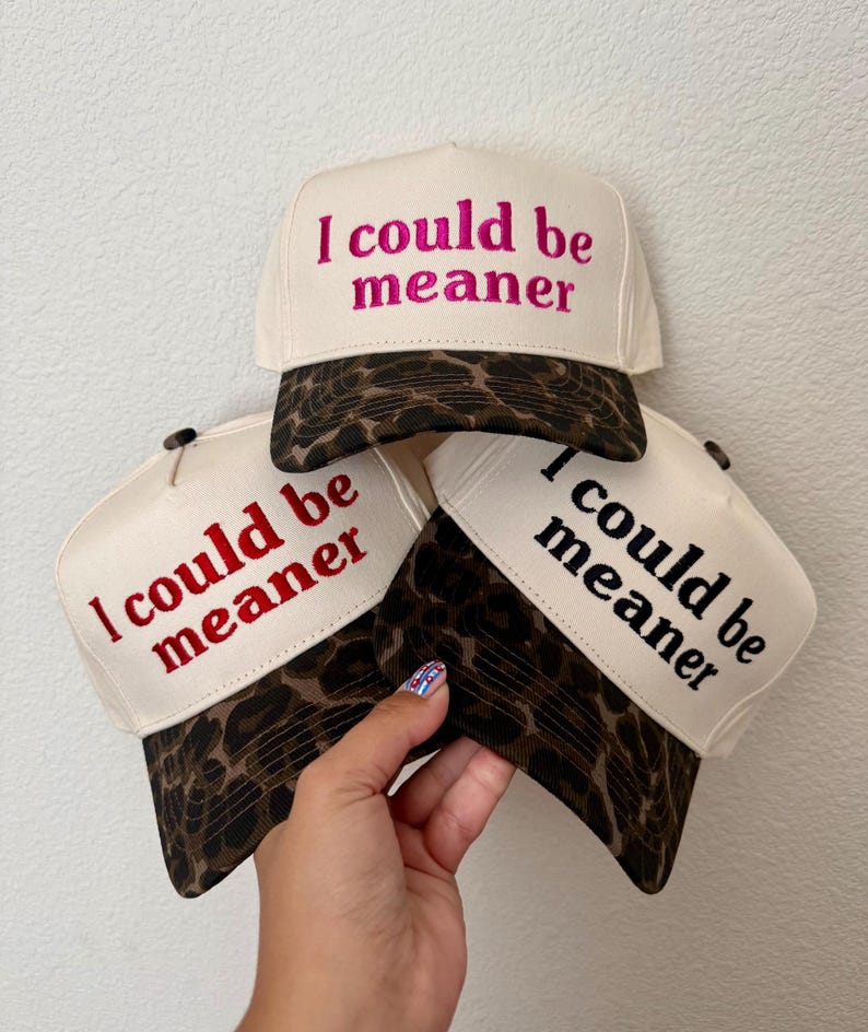 May include: Three trucker hats with cream-colored front panels and brown leopard print brims. Each hat features the embroidered text "I could be meaner" in different colors: one in hot pink, one in red, and one in black, displayed in a stacked arrangement.