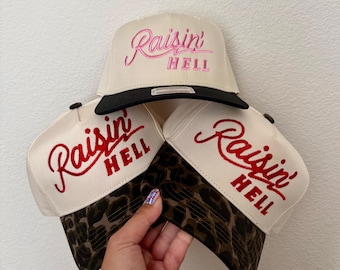 May include: Three baseball caps with off-white crowns and embroidered text that reads Raisin' HELL. One cap features a solid black brim, while the other two caps have brown and black leopard print brims. Each cap has a button on the top center.