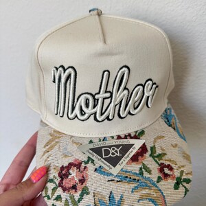 Puede incluir: Gorra de b&eacute;isbol color crema con la palabra "Mother" bordada en blanco y negro. La visera presenta un dise&ntilde;o de tapiz floral con detalles en rosa, azul y verde. Una etiqueta "D&Y" est&aacute; adherida a la visera.