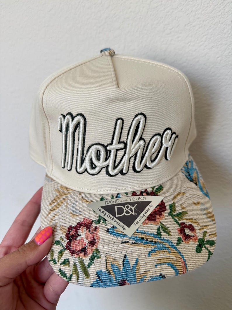Puede incluir: Gorra de b&eacute;isbol color crema con la palabra "Mother" bordada en blanco y negro. La visera presenta un dise&ntilde;o de tapiz floral con detalles en rosa, azul y verde. Una etiqueta "D&Y" est&aacute; adherida a la visera.