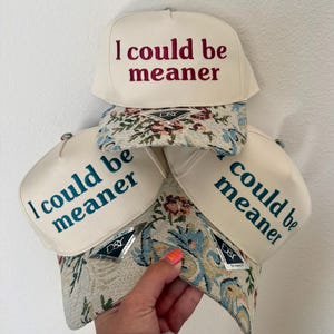 May include: Cream-colored baseball caps with the embroidered phrase "I could be meaner" in various colors. The caps have a floral patterned brim and a D&S tag.