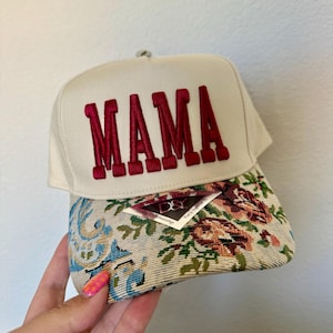 Puede incluir: Gorra de b&eacute;isbol color crema con bordado "MAMA" en borgo&ntilde;a. La visera presenta un dise&ntilde;o de tapiz floral con tonos azules, verdes y marrones. Una etiqueta est&aacute; adherida a la visera.