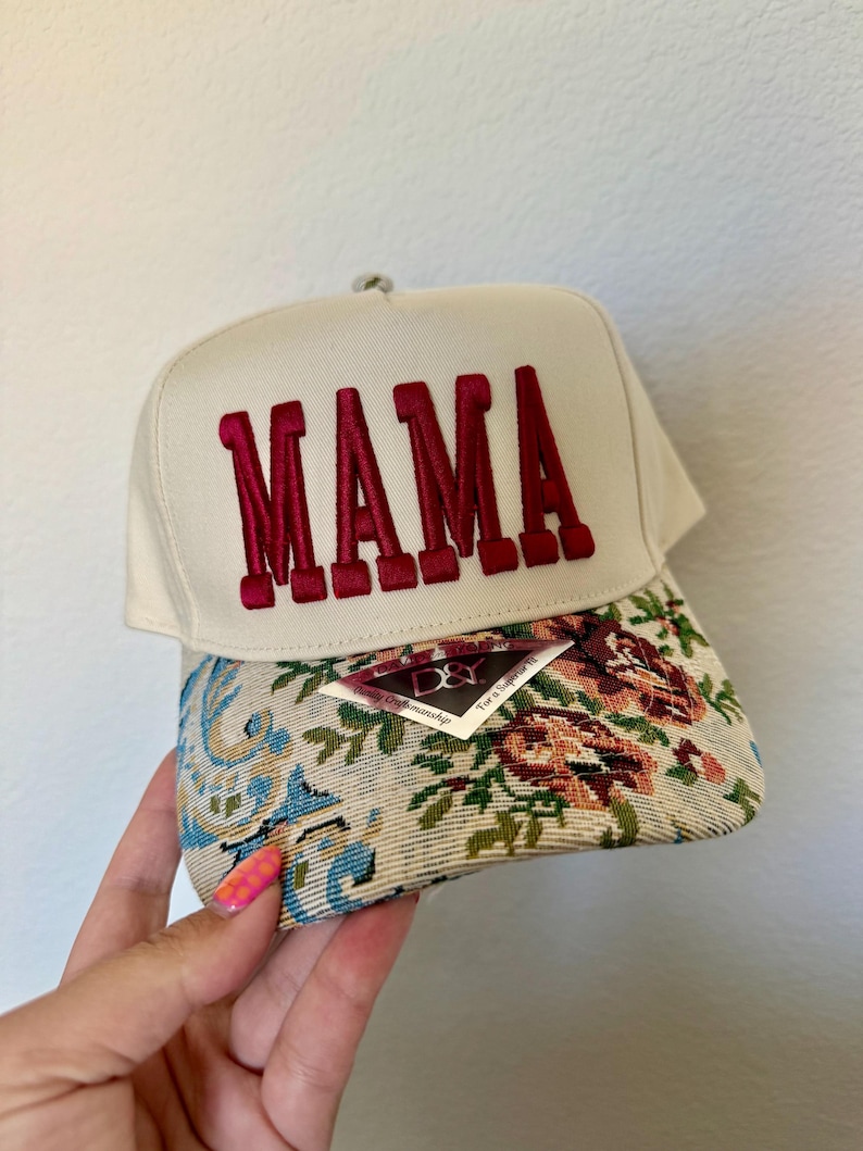 Puede incluir: Gorra de b&eacute;isbol color crema con bordado "MAMA" en borgo&ntilde;a. La visera presenta un dise&ntilde;o de tapiz floral con tonos azules, verdes y marrones. Una etiqueta est&aacute; adherida a la visera.