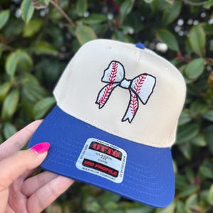 May include: A beige and blue baseball cap with a baseball-themed bow design. The cap features a blue brim and a beige crown with a red and white embroidered bow. The cap has a "OTTO" label.