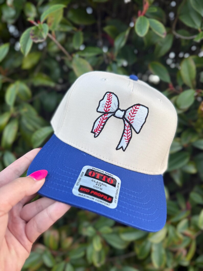 May include: A beige and blue baseball cap with a baseball-themed bow design. The cap features a blue brim and a beige crown with a red and white embroidered bow. The cap has a "OTTO" label.