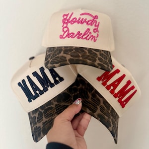 May include: Three cream-colored baseball caps with leopard print brims. The top cap features pink embroidered text reading Howdy Darlin'. The bottom left cap has navy blue embroidered text reading MAMA, and the bottom right cap has red text.