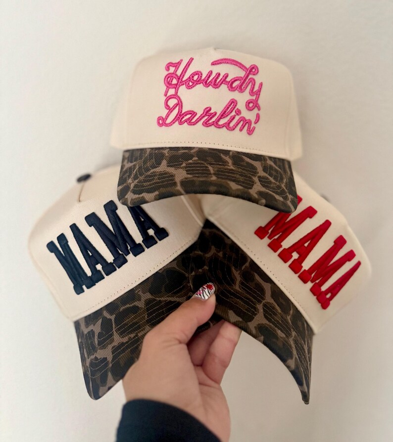 May include: Three cream-colored baseball caps with leopard print brims. The top cap features pink embroidered text reading Howdy Darlin'. The bottom left cap has navy blue embroidered text reading MAMA, and the bottom right cap has red text.