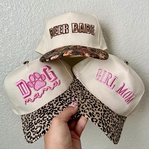 May include: Three beige baseball caps with different embroidered text and patterned brims. One cap says "BEER BABE" with a camouflage brim. Another says "DOG mama" with a pink paw print and a leopard print brim. The third says "GIRL MOM" with a leopard print brim.