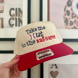 May include: A baseball cap with a cream-colored crown and a red brim. The cap features embroidered text that reads "Take me out to the Ball Game" with a baseball graphic. The cap is being held by a person.