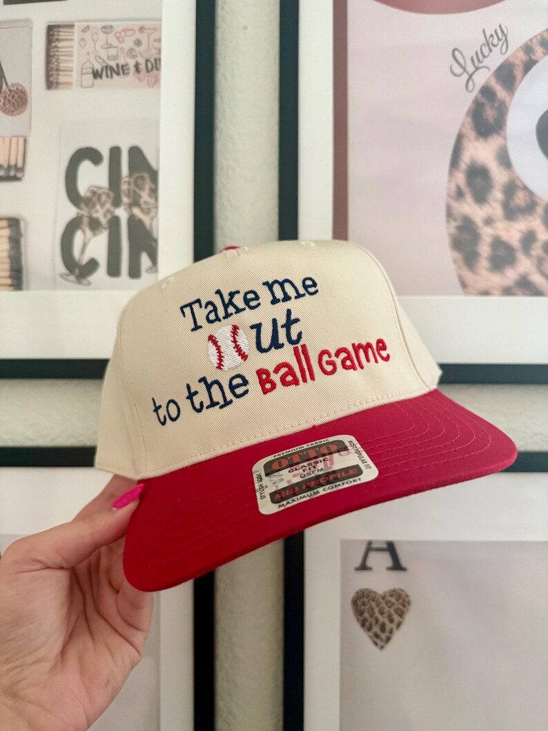 May include: A baseball cap with a cream-colored crown and a red brim. The cap features embroidered text that reads "Take me out to the Ball Game" with a baseball graphic. The cap is being held by a person.