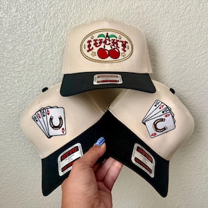 May include: Three beige and black trucker hats. One hat features the word "LUCKY" with cherries. The other two hats have playing card and horseshoe designs. Each hat has a black brim and a patch.
