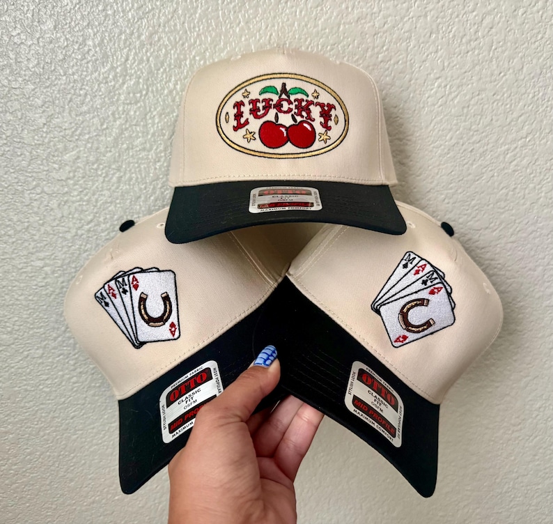 May include: Three beige and black trucker hats. One hat features the word "LUCKY" with cherries. The other two hats have playing card and horseshoe designs. Each hat has a black brim and a patch.