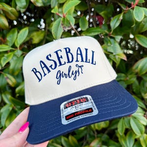May include: A baseball cap with a cream-colored crown and a navy blue brim. The words "BASEBALL girly" are embroidered in navy blue script on the front. The cap has a classic fit and a mid-profile design.