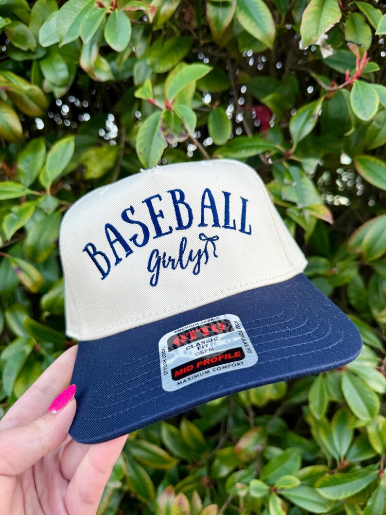 May include: A baseball cap with a cream-colored crown and a navy blue brim. The words "BASEBALL girly" are embroidered in navy blue script on the front. The cap has a classic fit and a mid-profile design.