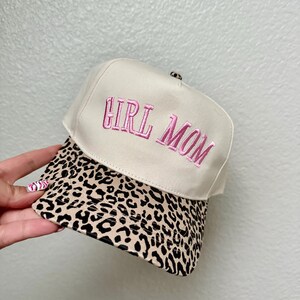 May include: A beige baseball cap with the embroidered text "GIRL MOM" in pink. The brim features a leopard print design. The hat is a stylish accessory.