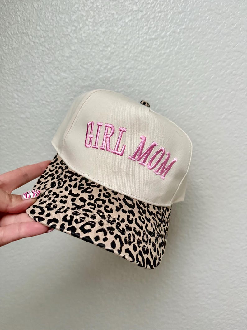 May include: A beige baseball cap with the embroidered text "GIRL MOM" in pink. The brim features a leopard print design. The hat is a stylish accessory.
