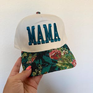 Puede incluir: Gorra de b&eacute;isbol blanca con la palabra "MAMA" bordada en color turquesa. La visera presenta un estampado floral con detalles en rojo, verde y negro. La gorra est&aacute; sujeta por una mano.