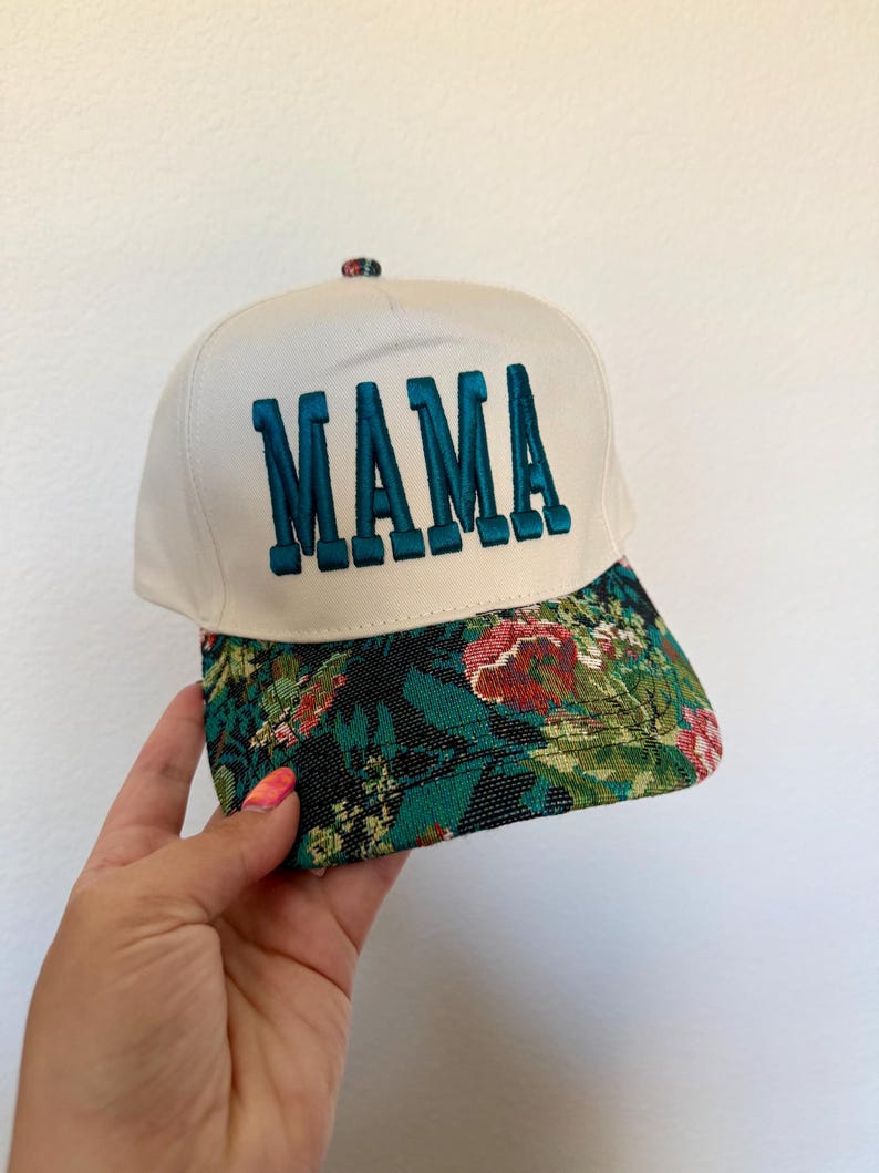 Puede incluir: Gorra de b&eacute;isbol blanca con la palabra "MAMA" bordada en color turquesa. La visera presenta un estampado floral con detalles en rojo, verde y negro. La gorra est&aacute; sujeta por una mano.