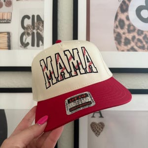 May include: A baseball cap with a cream-colored crown and a red brim. The word "MAMA" is embroidered in red with baseball stitching. The cap has a curved brim and a red button on top.