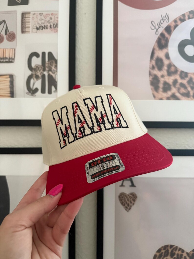 May include: A baseball cap with a cream-colored crown and a red brim. The word "MAMA" is embroidered in red with baseball stitching. The cap has a curved brim and a red button on top.