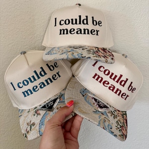 May include: Off-white baseball caps with the embroidered text "I could be meaner" in black, teal, and red. The caps have patterned brims with floral and tapestry designs. The caps are stacked together.