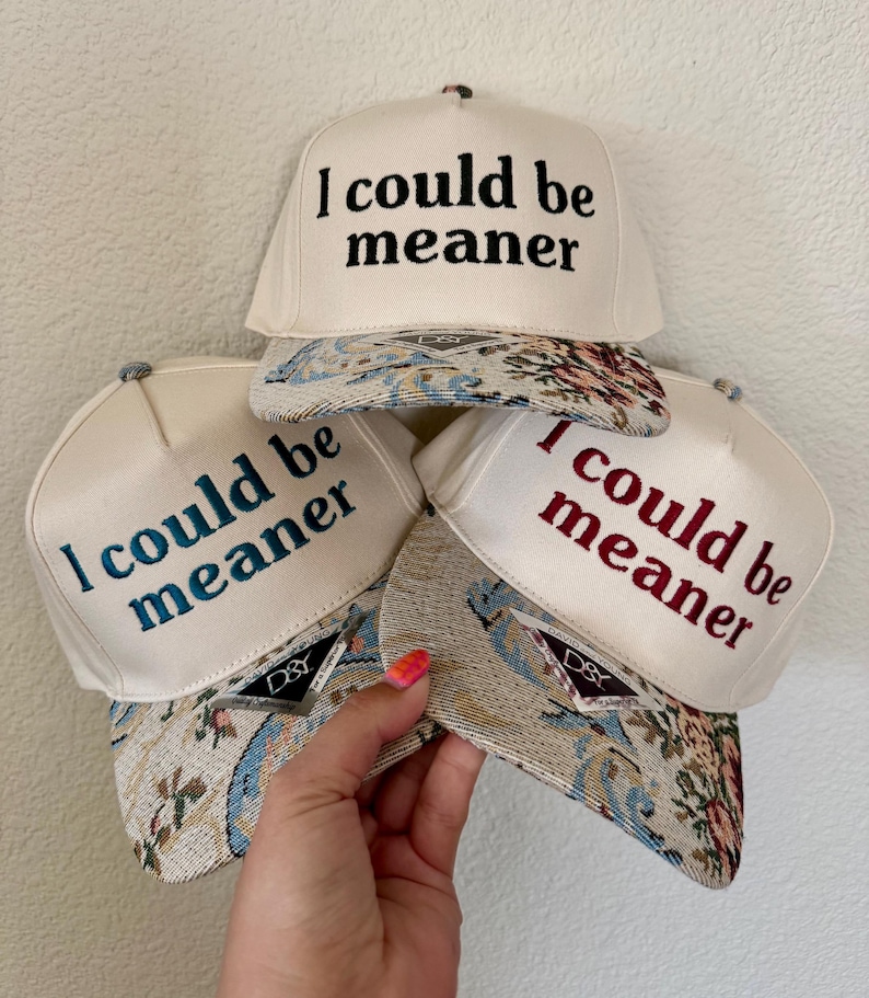May include: Off-white baseball caps with the embroidered text "I could be meaner" in black, teal, and red. The caps have patterned brims with floral and tapestry designs. The caps are stacked together.