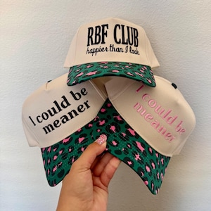 May include: Three beige baseball caps with black and pink text and a green and pink leopard print brim. One cap reads "RBF CLUB happier than I look". The other two caps read "I could be meaner".