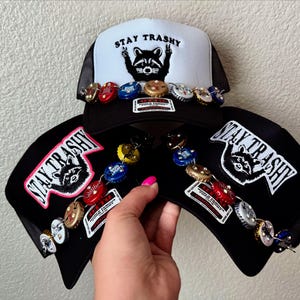 May include: Three black trucker hats with the words "Stay Trashy" embroidered on them. Each hat has a raccoon graphic and is decorated with bottle caps.
