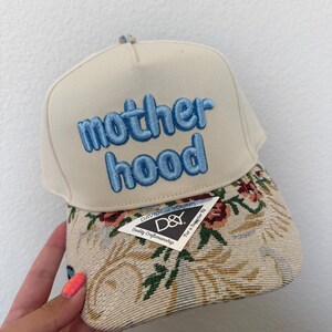 Puede incluir: Gorra de b&eacute;isbol beige con la palabra "motherhood" bordada en azul. La visera presenta un dise&ntilde;o de tapiz floral y una etiqueta "David & Young".